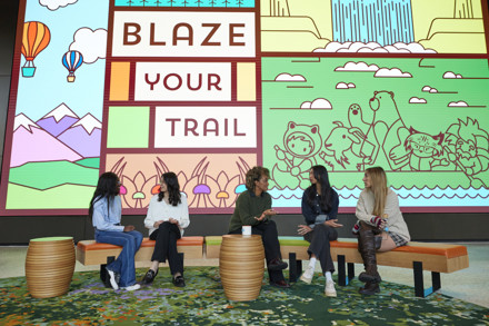 Five women are seated on benches in front of a colorful digital backdrop that says "Blaze Your Trail" with illustrations of Salesforce mascots.