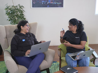 Two Salesforce employees are having a conversation in an office, they both holding their laptops on their laps.