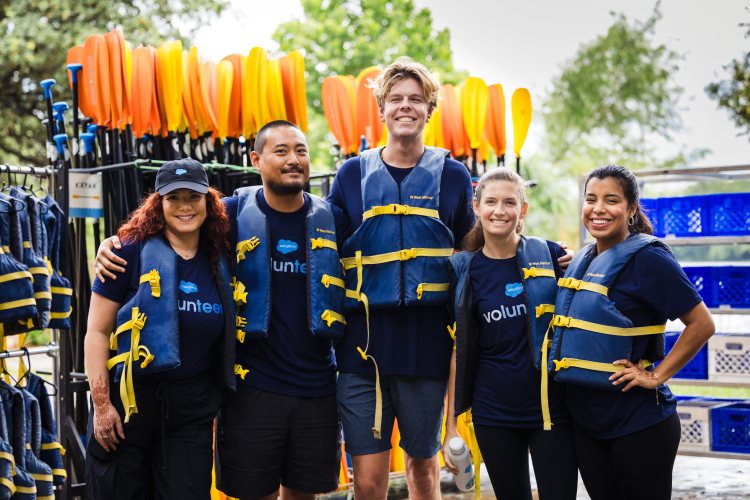 A group of Salesforce volunteers wearing life vests pose together in front of kayaks, ready for an outdoor team activity.