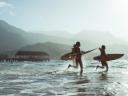 Three people on a beach, running into the ocean with surboards in a tropical climate, with mountains in the background.