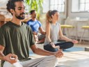 A man with a ponytail meditates in a yoga class, surrounded by other yoga class participants in a bright studio with large windows and plants.