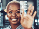 A black woman with close-cropped blond hair is tapping or swiping at data that's displayed on a clear screen in the foreground.