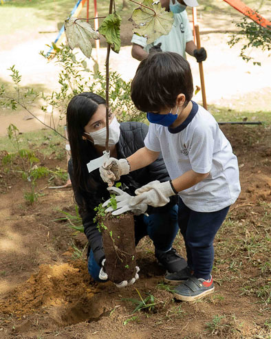 Beatriz knees on the ground, holding a tree with a young boy as they plant it into the ground together.