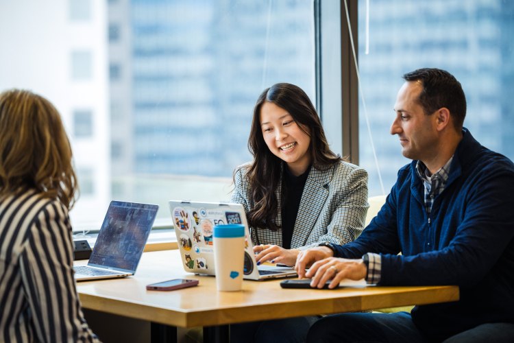 A group of Salesforce employees around a small table, with laptops open and Salesforce mugs surrounding them. Behind them, a wall of windows shows the Indianapolis skyline.