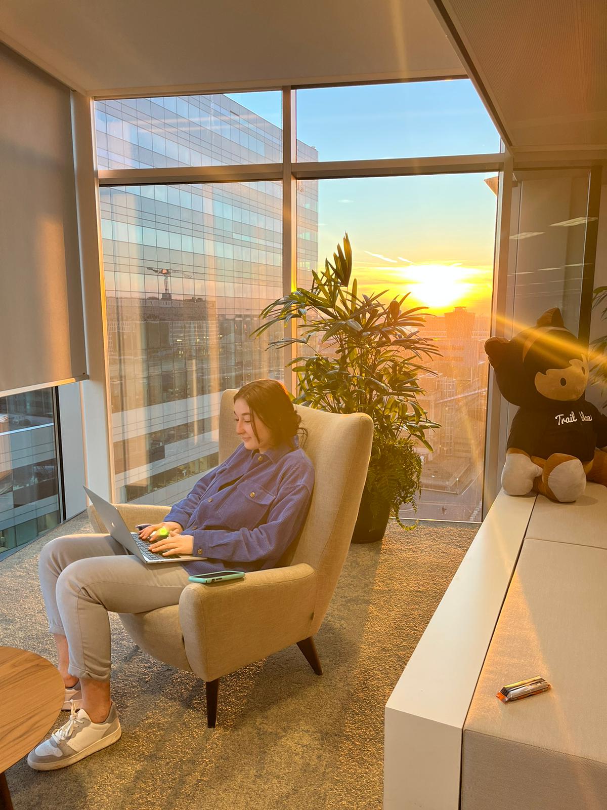 Kelsey sits in a lounge chair in the Amsterdam office with an Astro plush on the desk beside her. The sunlight casts a gold glow over the room.