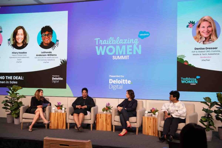 A panel of four women sit on a stage under a screen displaying the Trailblazing Women Summit logo