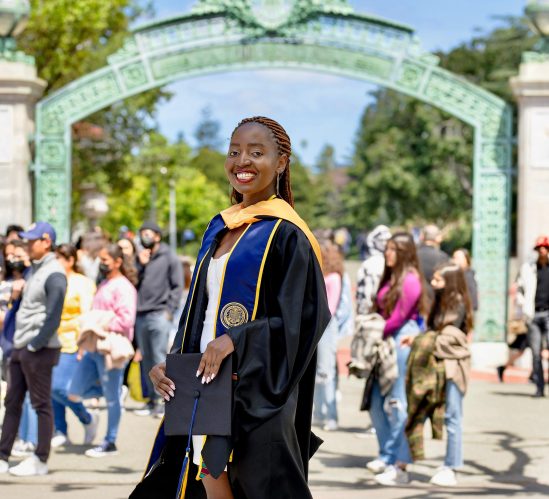 Gloria Tumushabe in her graduation cap and gown in front of a green metal archway