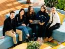 A group of five young professionals sits together on a blue couch, smiling and collaborating around a laptop in a bright, modern office space.