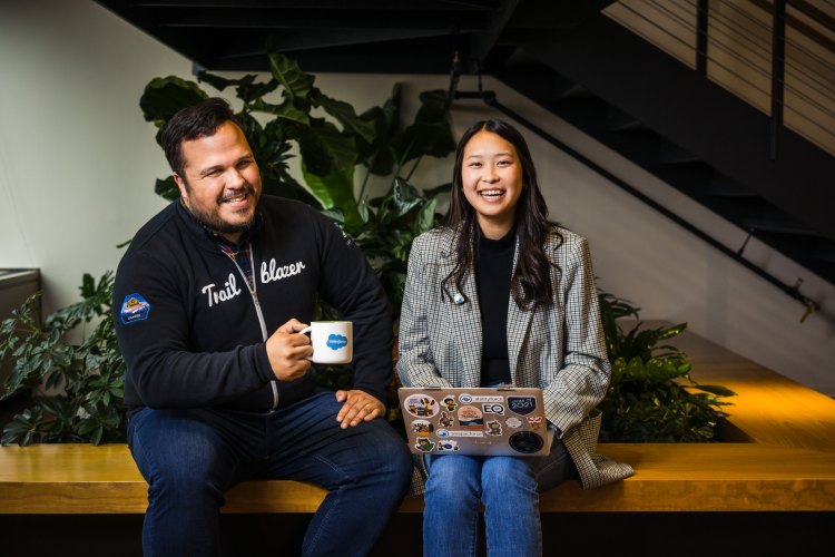 Two Salesforce employees sit next to each other, smiling. One holds a coffee mug and the other holds a laptop.