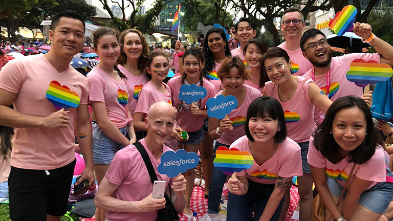 A group of Salesforce Singapore employees attend the Pride Month parade, wearing pink t-shirts and holding rainbow-colored cloud-shaped figures.
