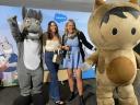 Flavia and another Salesforce employee are smiling while standing next to Salesforce mascots, Max and Astro.