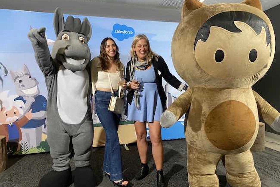 Flavia and another Salesforce employee are smiling while standing next to Salesforce mascots, Max and Astro.
