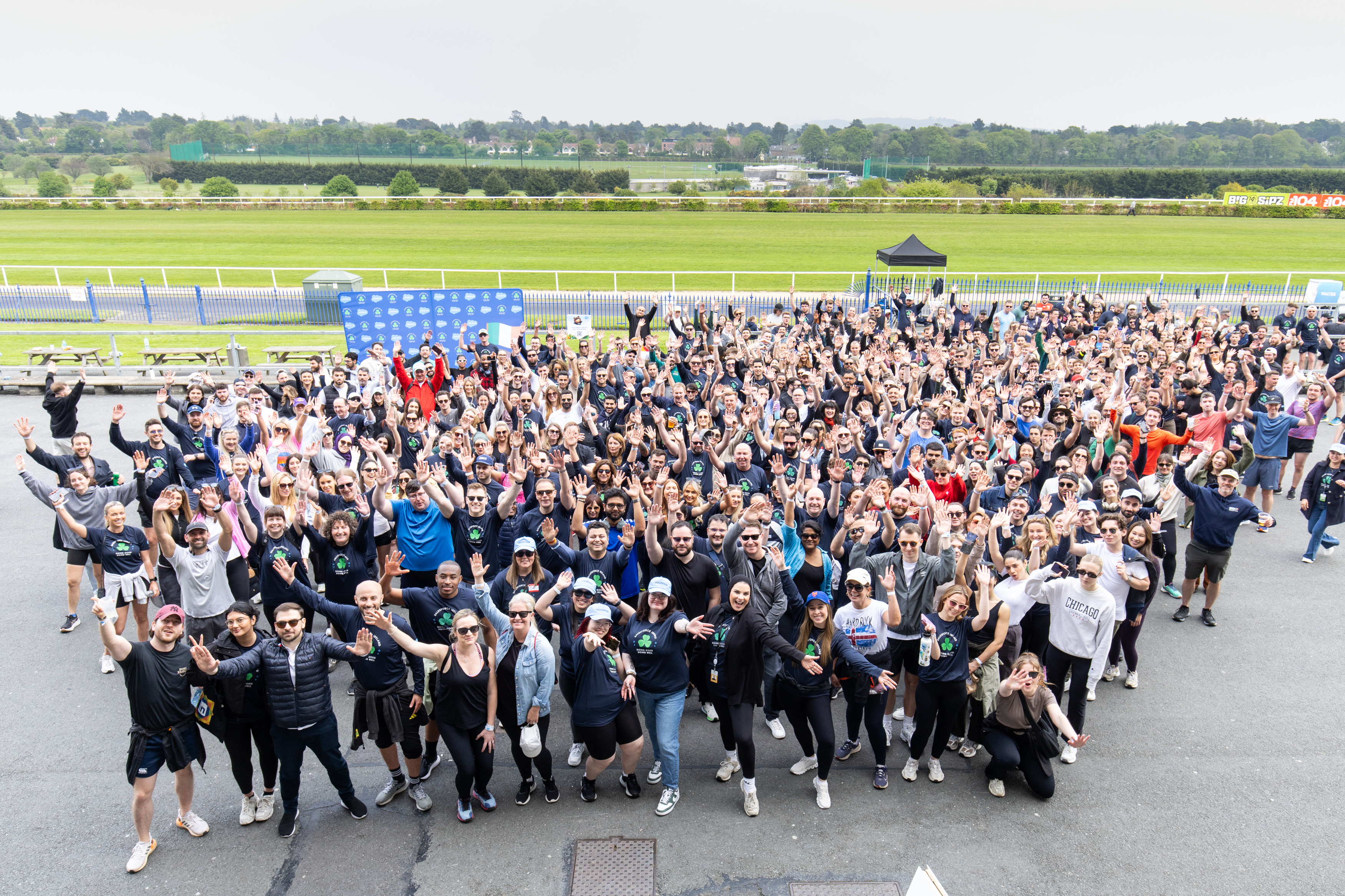 A large group of people is gathered outdoors, smiling and waving at the camera, suggesting a celebratory or team-building event.
