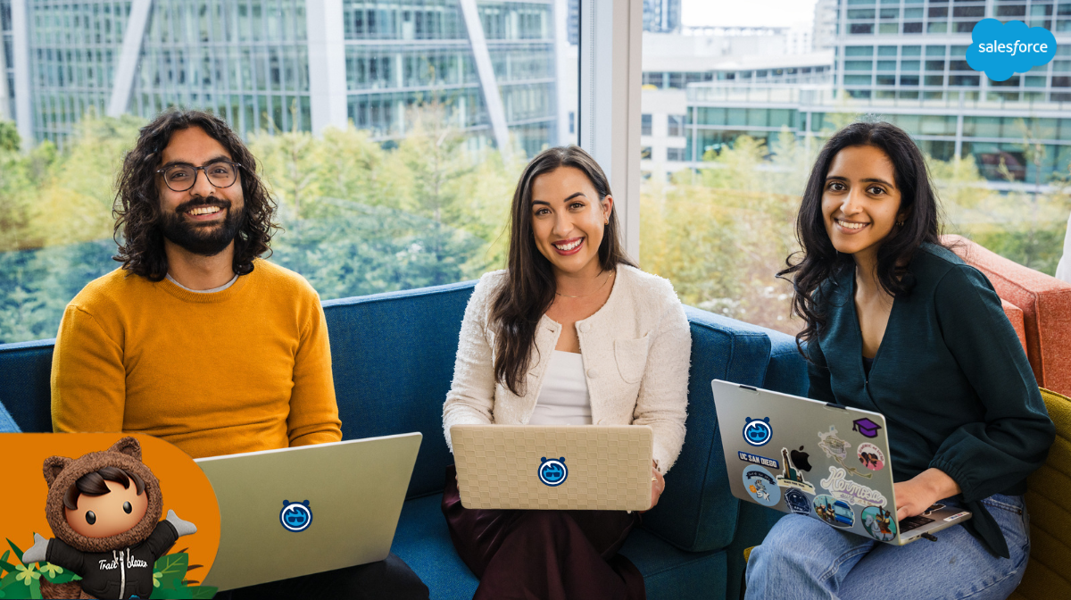 three employees sit in a modern office setting and pose with their computers