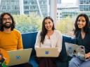 three employees sit in a modern office setting and pose with their computers