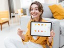 A woman sitting in a living room with yellow pillows and an online store tablet, pointing at it, smiling.