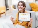 A woman sitting in a living room with yellow pillows and an online store tablet, pointing at it, smiling.