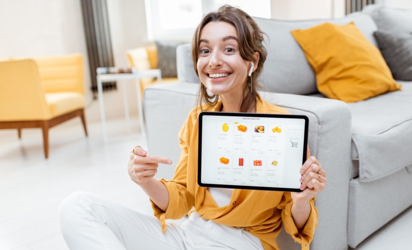 A woman sitting in a living room with yellow pillows and an online store tablet, pointing at it, smiling.
