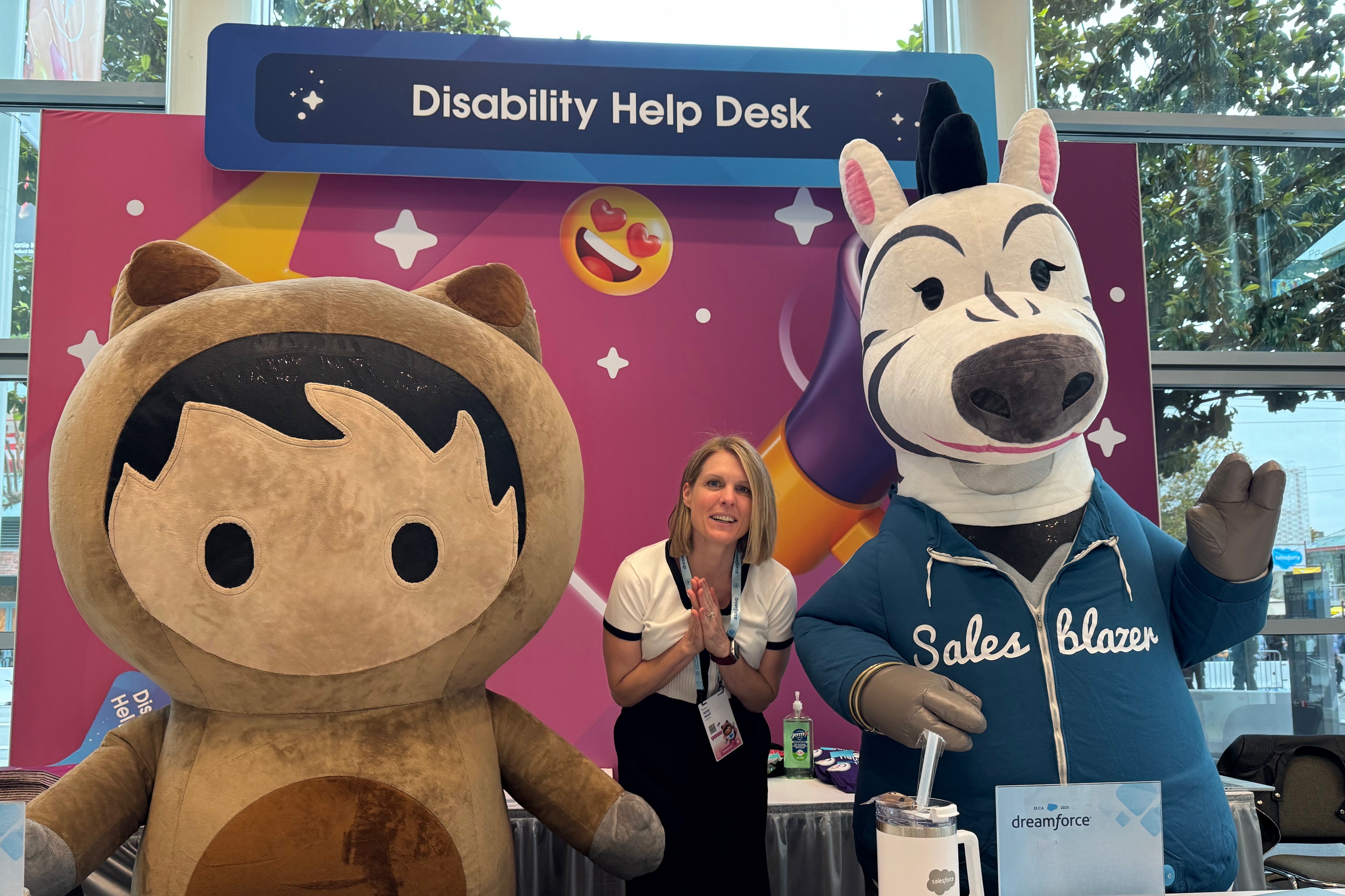 A blonde woman stands between two Salesforce mascots: Astro, a bear-like mascot, and Ziggy, a zebra mascot wearing a Salesblazer Hoodie. They pose for the photo in front of the pink and blue Disability Help Desk sign decorated with stars and a smiling emoji with heart eyes.