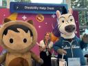A blonde woman stands between two Salesforce mascots: Astro, a bear-like mascot, and Ziggy, a zebra mascot wearing a Salesblazer Hoodie. They pose for the photo in front of the pink and blue Disability Help Desk sign decorated with stars and a smiling emoji with heart eyes.
