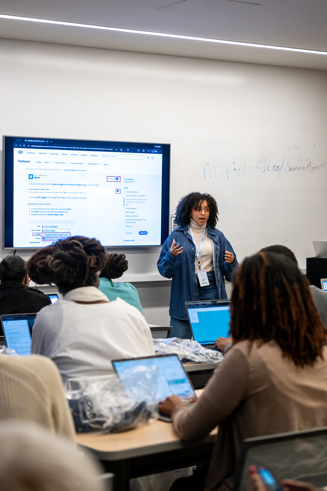 Rosa Moss of Salesforce, in a blue denim shacket and white turtleneck, stands at the front of a filled classroom presenting to an audience of attendees with their laptops open. Behind her, the screen shows a demo Service Cloud instance.