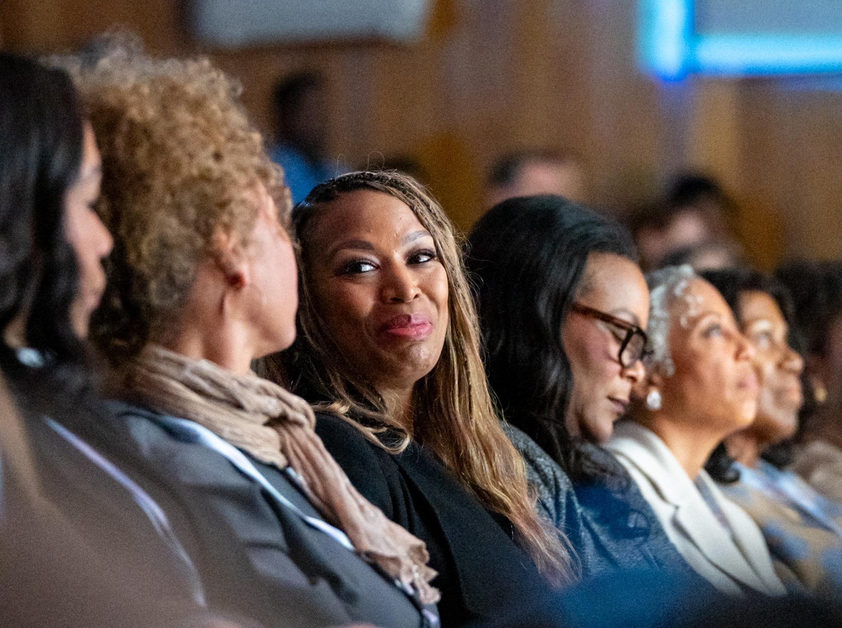 A row of women sit in auditorium-style seating, facing the stage. Two are engaging with each other, smiling.