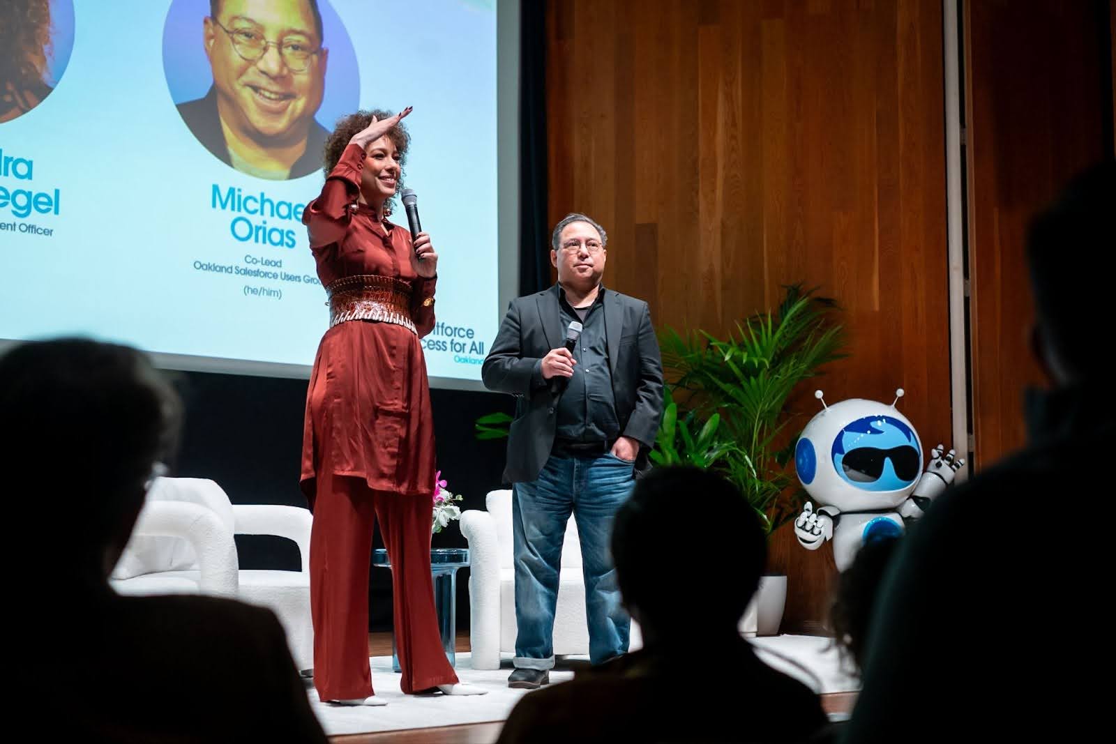 Alexandra Siegel in a matching maroon outfit and Michael Orias in a black jacket and shirt and jeans, stand together onstage and look out to the audience. Behind them, wood paneling, white club chairs, a giant Agent Astro mascot, and a display screen with both speakers' headshots and titles.