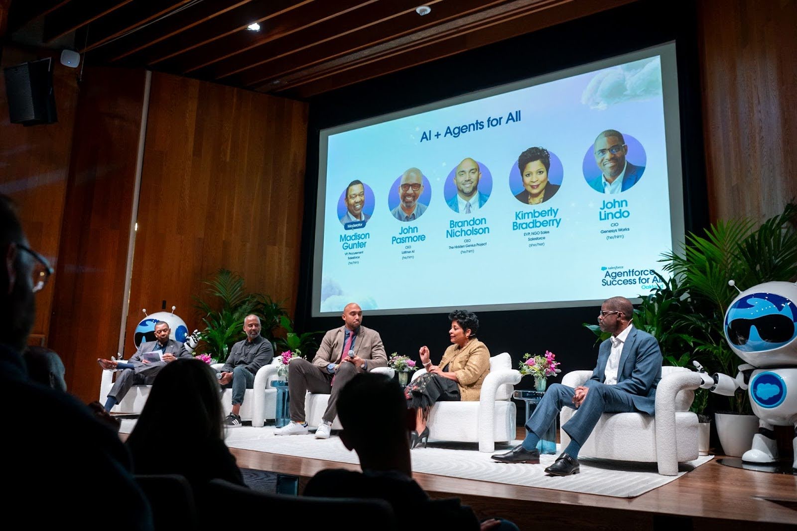 Panelists sit onstage in white club chairs. Behind them, warm wood walls and a giant screen showcasing each speaker's headshot and title. A giant Salesforce Agent Astro mascot stands to the right of the speakers.