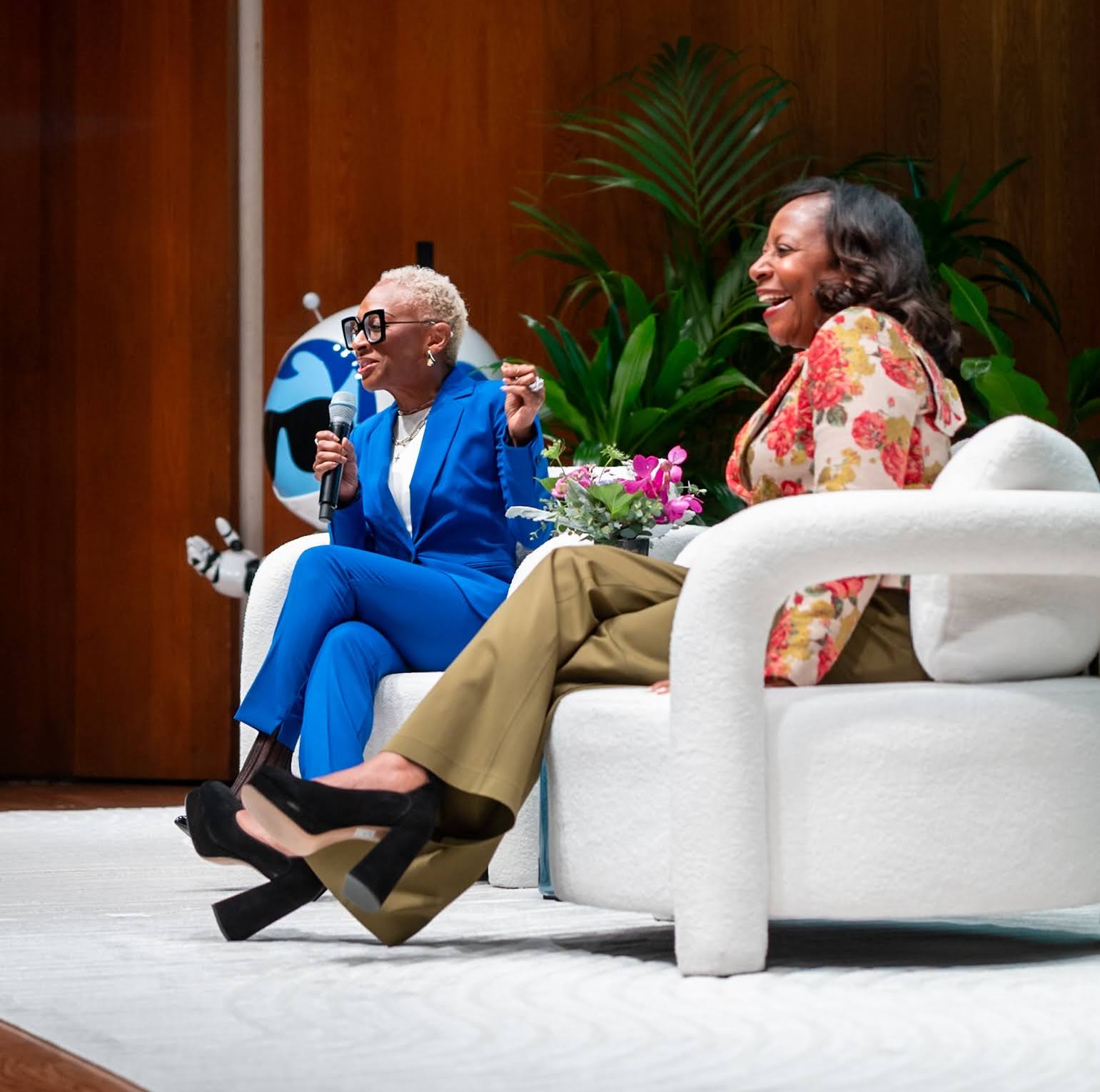 LaShonda Anderson-Williams in a blue suit and Robin Washington in a floral top and dark khaki pants sit in white club chairs onstage, with warm wood walls, greenery and a giant Salesforce Astro mascot behind them.