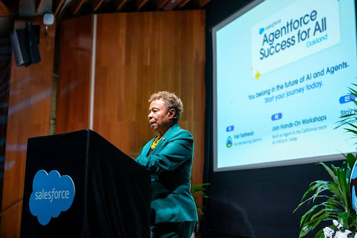 Oakland Mayor Barbara Lee in a turquoise suit at a podium with the Salesforce cloud logo in the center; behind her, the screen displays the event logo, "Agentforce Success for All Oakland" with light and dark blues.