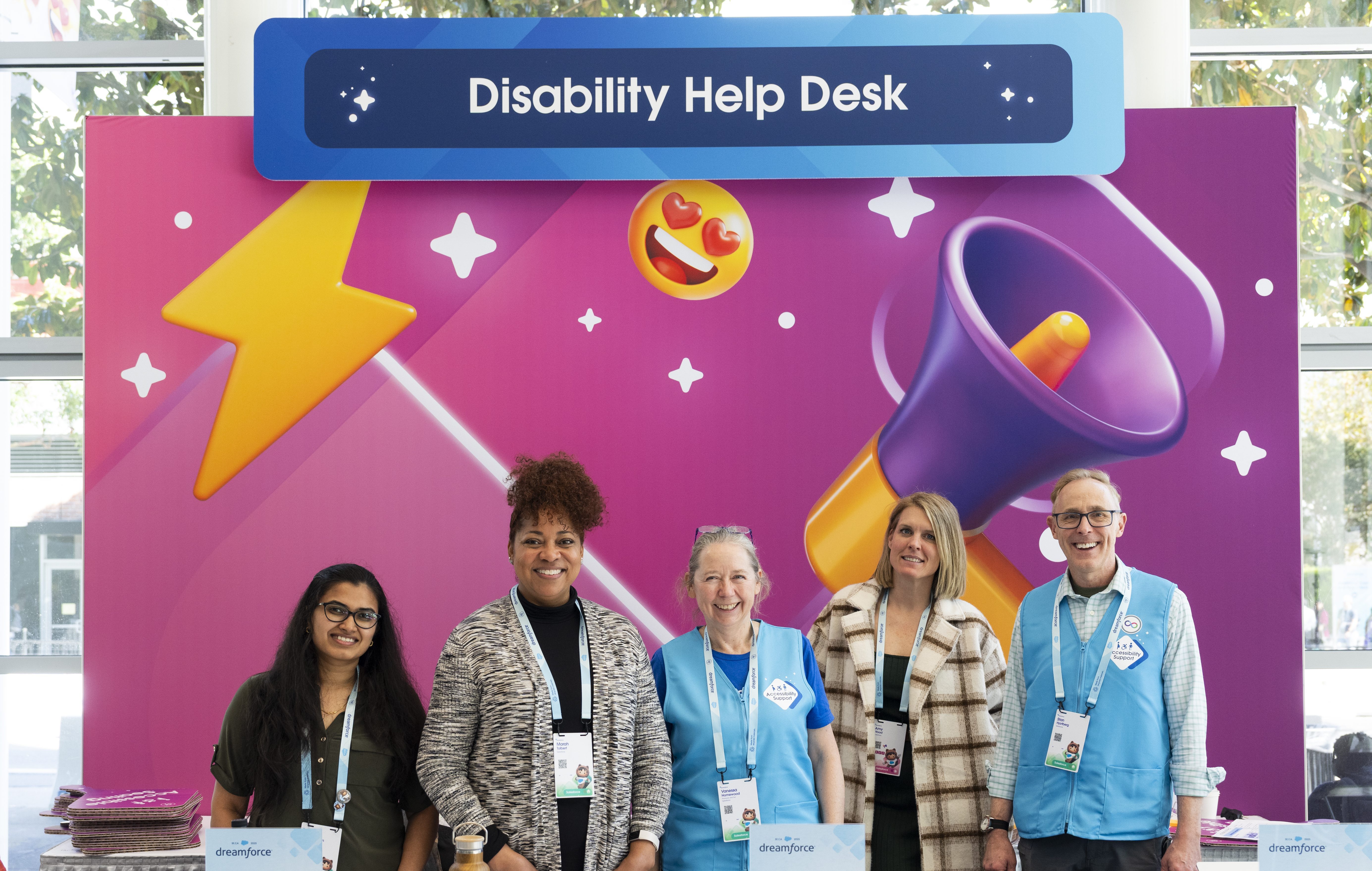 A group of five people stand behind a desk. Two wear bright blue vests marking them as Accessibility Guides. Behind them, a large backdrop in Salesforce's pink Dreamforce branding with yellow and purple emojis. The top of the background has a navy header plaque that displays "Disability Help Desk"