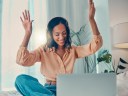 A woman with headphones on and a laptop working on guest blogging on a comfortable bed with her arms raised in celebration.