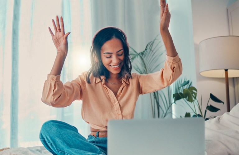 A woman with headphones on and a laptop working on guest blogging on a comfortable bed with her arms raised in celebration.