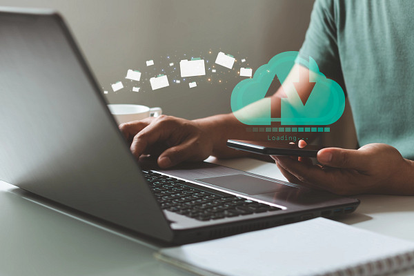 An illustration of cloud computing showing a person syncing data between a laptop and a smartphone, represented by a glowing cloud icon, a loading bar, and floating file folders.