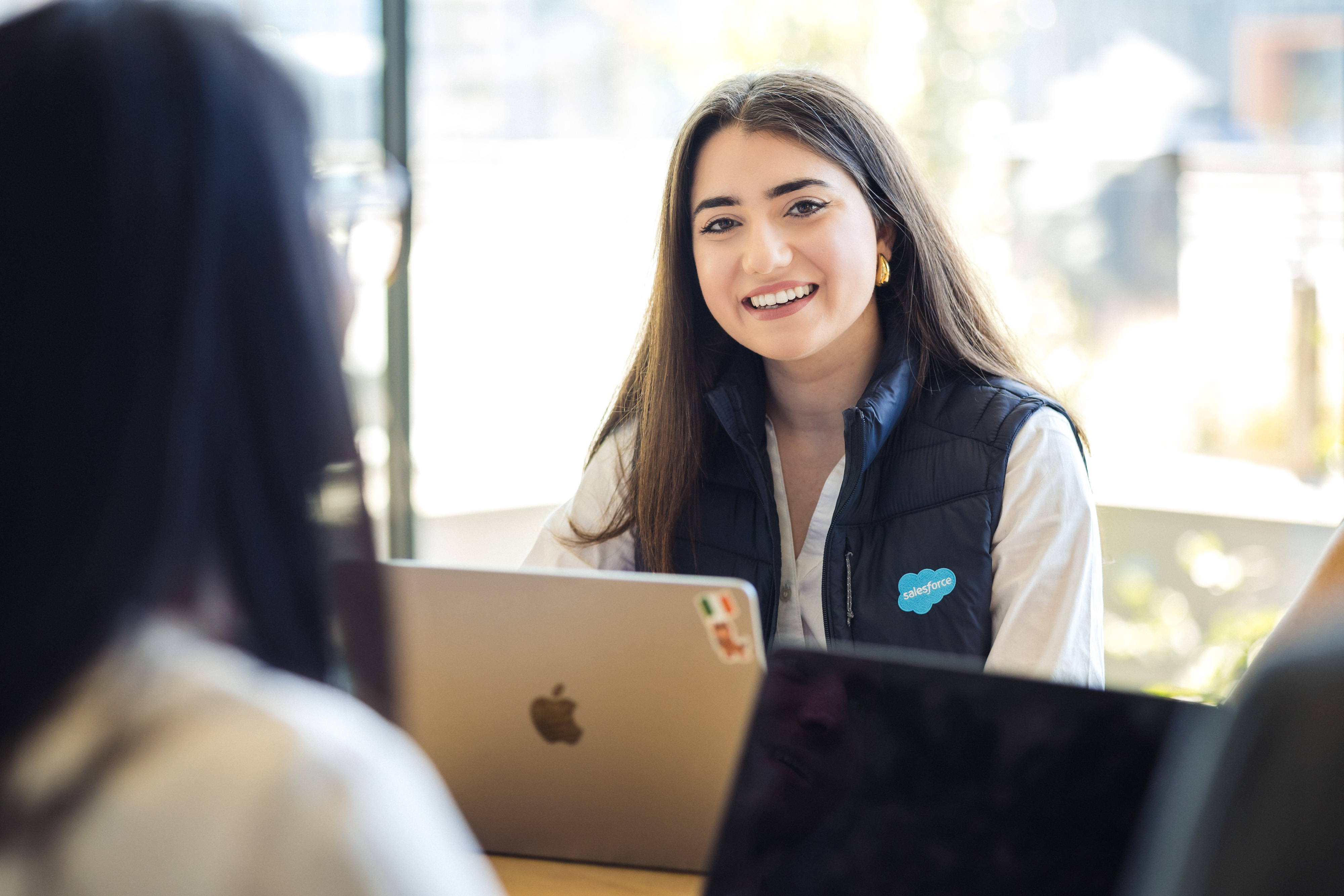 A smiling person wearing a navy Salesforce vest sits at a table with a laptop, looking toward the camera during a conversation with a colleague.