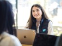 A smiling person wearing a navy Salesforce vest sits at a table with a laptop, looking toward the camera during a conversation with a colleague.