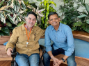Alfonso, a Workforce Navigators participant, sits in his power wheelchair next to his Salesforce mentor, Saurabh, who is seated on a bench. They are smiling together in a lush, indoor garden setting at Salesforce Tower.