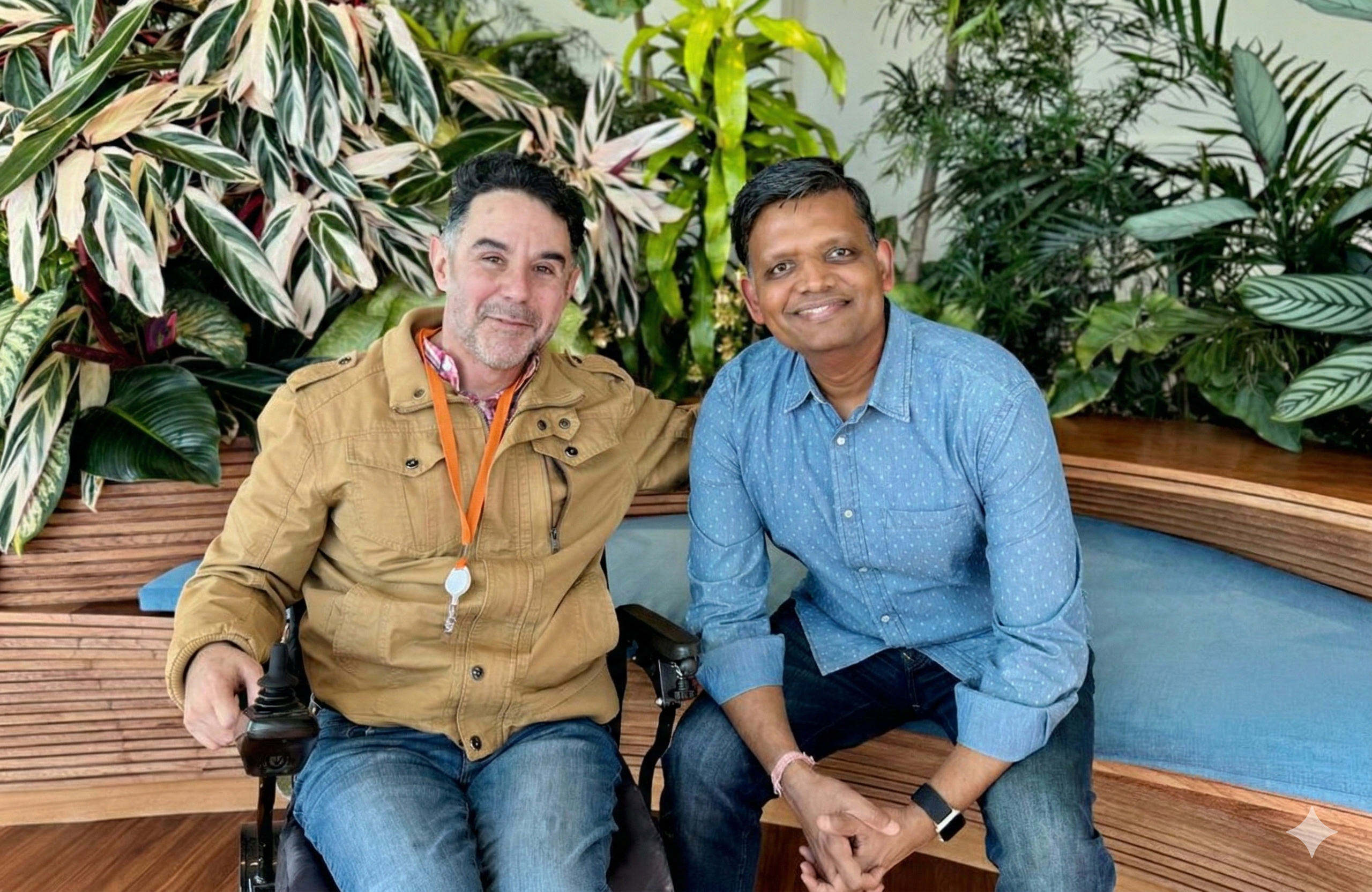 Alfonso, a Workforce Navigators participant, sits in his power wheelchair next to his Salesforce mentor, Saurabh, who is seated on a bench. They are smiling together in a lush, indoor garden setting at Salesforce Tower.