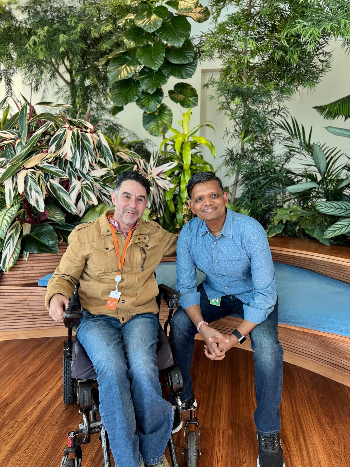 Alfonso, a Workforce Navigators participant, sits in his power wheelchair next to his Salesforce mentor, Saurabh, who is seated on a bench. They are smiling together in a lush, indoor garden setting at Salesforce Tower.