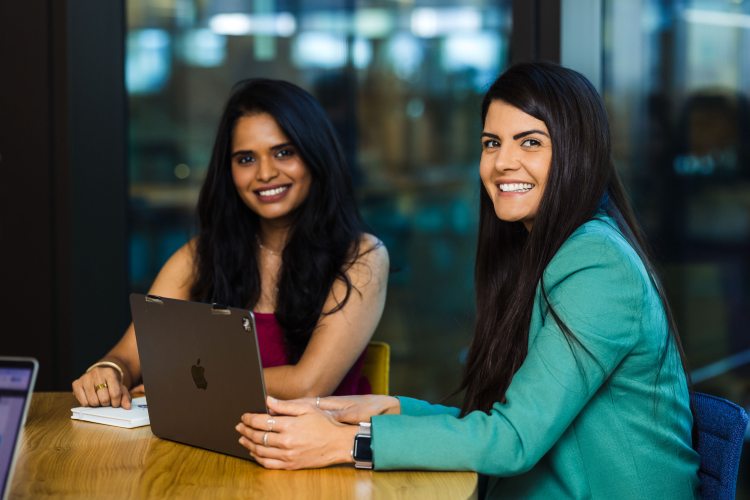 One person smiles at the camera while working on a laptop, another at the desk smiles while looking at the laptop.