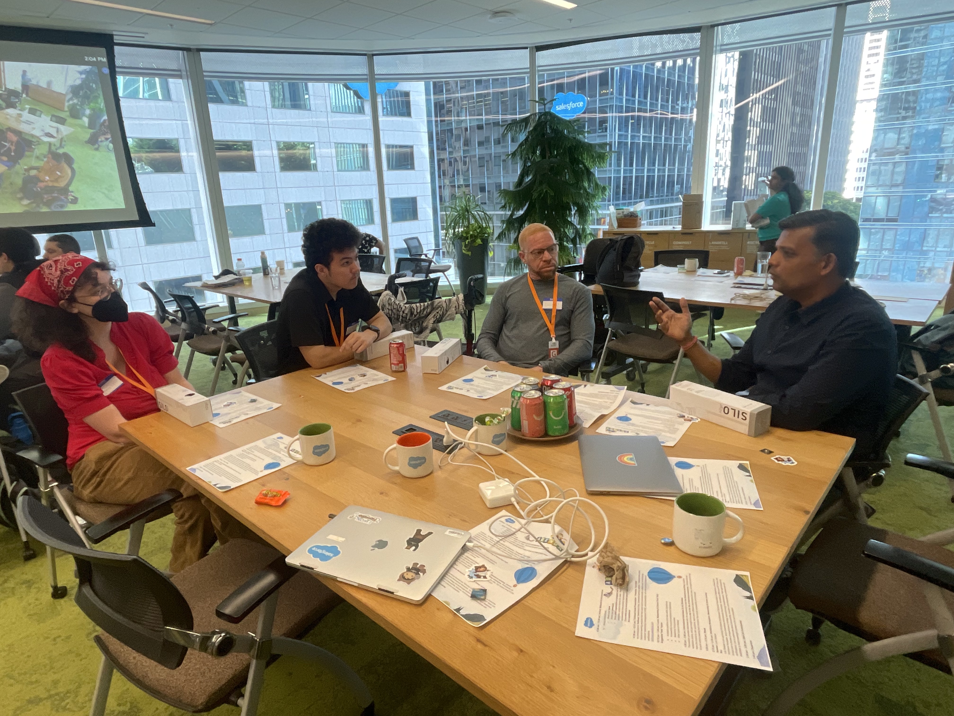 Four participants sit around a wooden table in a bright office space with floor-to-ceiling windows overlooking San Francisco. One person on the right gestures while speaking to the group. The table is covered with Salesforce-branded mugs, laptops, and workshop handouts.