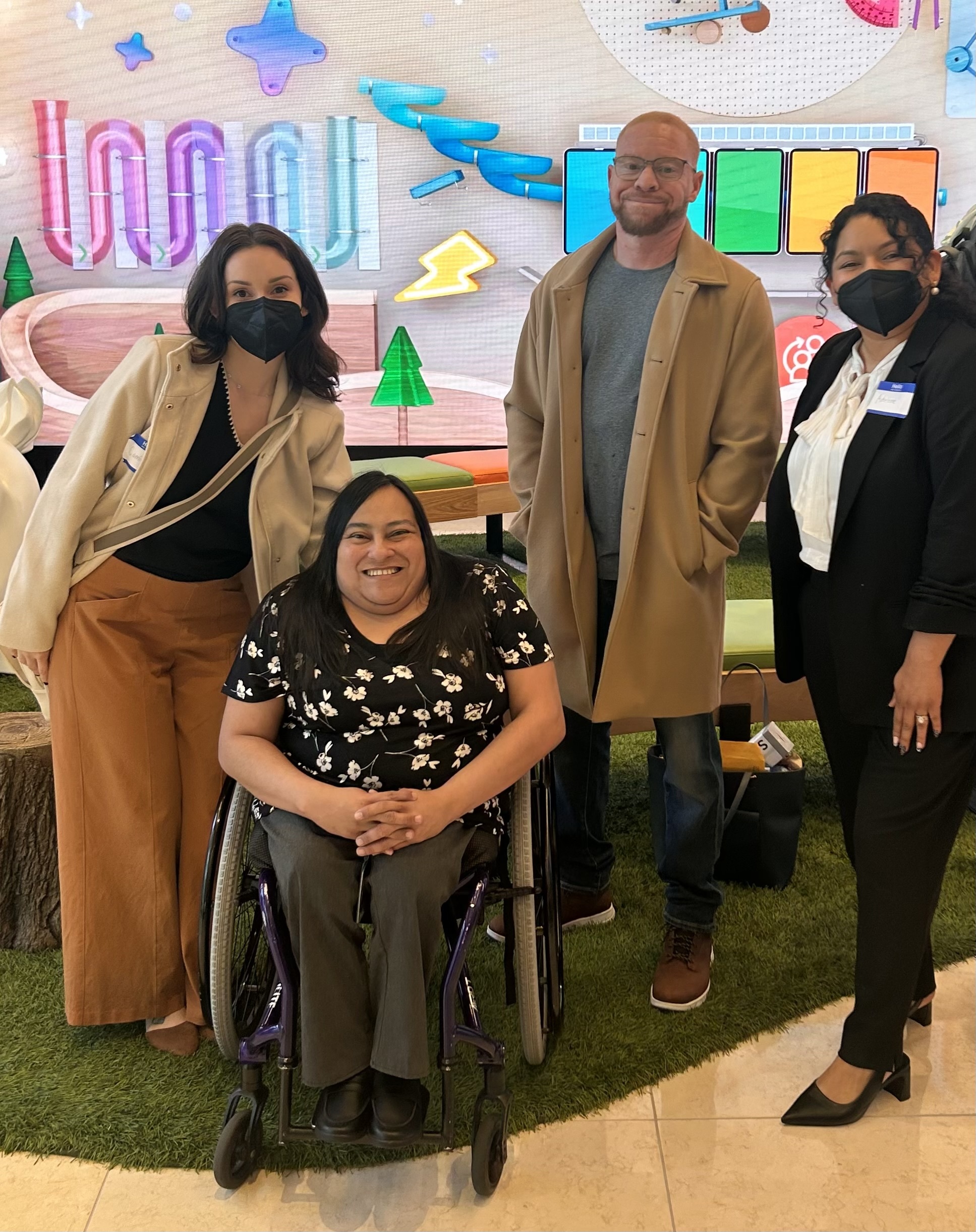 A group photo showing Camile Hayes (left) and Lucia Rios (second from left), who is in a wheelchair. Two Center for Independent Living staff members are to Rios's right. They stand on artificial turf in front of a modern, geometric-patterned wall with neon lights and digital screens in the lobby of the Salesforce San Francisco Tower.
