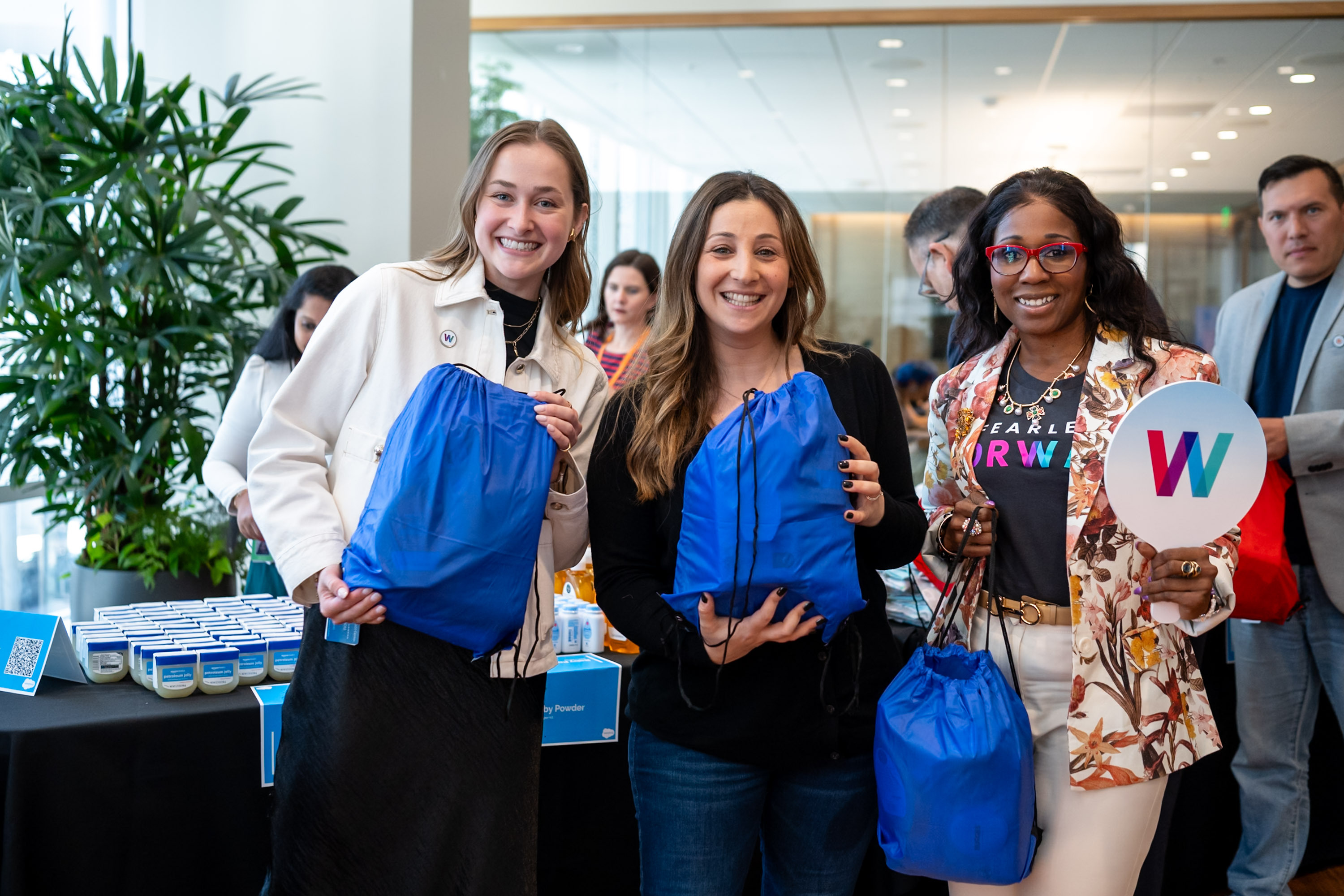 Three smiling attendees pose together at a Salesforce Women's Network event, each holding a blue drawstring swag bag. The person on the right also holds a circular sign featuring a colorful "W" logo.
