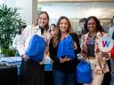 Three smiling attendees pose together at a Salesforce Women's Network event, each holding a blue drawstring swag bag. The person on the right also holds a circular sign featuring a colorful "W" logo.