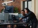 Two software developers holding laptop with coding interface walking towards desk and sitting down talking about group project.