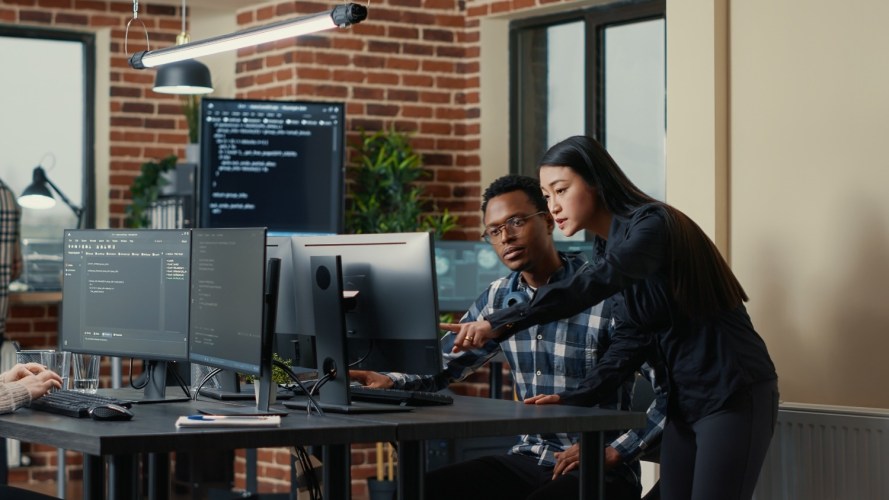 Two software developers holding laptop with coding interface walking towards desk and sitting down talking about group project.