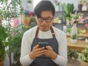 A young adult asian man in a florist shop looking at his phone surrounded by various indoor plants.