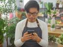 A young adult asian man in a florist shop looking at his phone surrounded by various indoor plants.