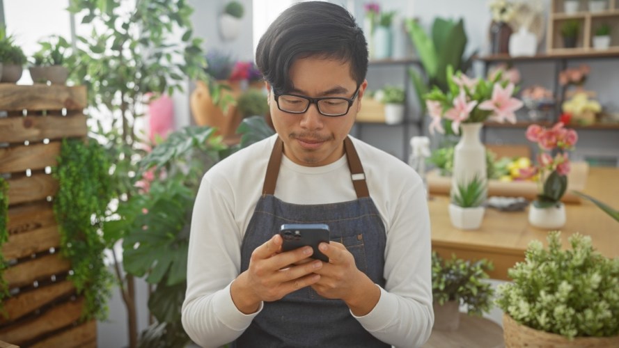 A young adult asian man in a florist shop looking at his phone surrounded by various indoor plants.