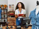 Young latin shopkeeper woman using touchpad working at clothing store.